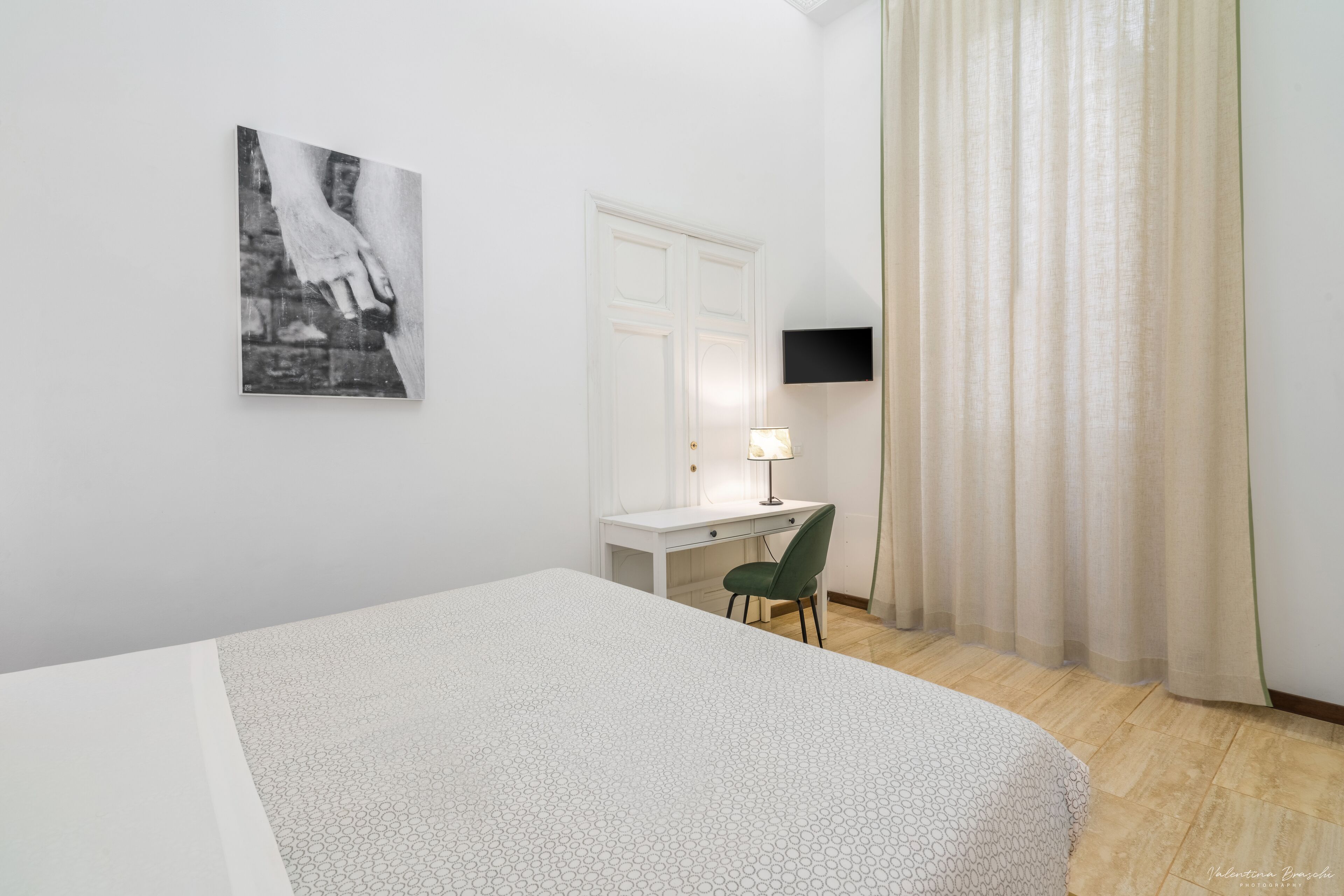 Un dormitorio con cama, escritorio, silla, televisión montada en la pared y ventana con cortinas.