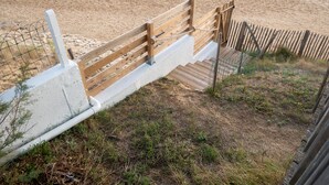 Un escalier en bois descend vers une plage de sable, et un garde-corps et une clôture bordent le chemin.