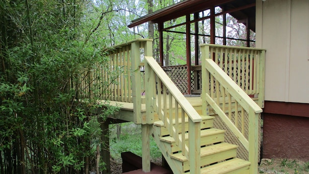 Un escalier en bois mène à une terrasse dotée d’une rambarde en treillis, le tout étant entouré d’une végétation luxuriante.
