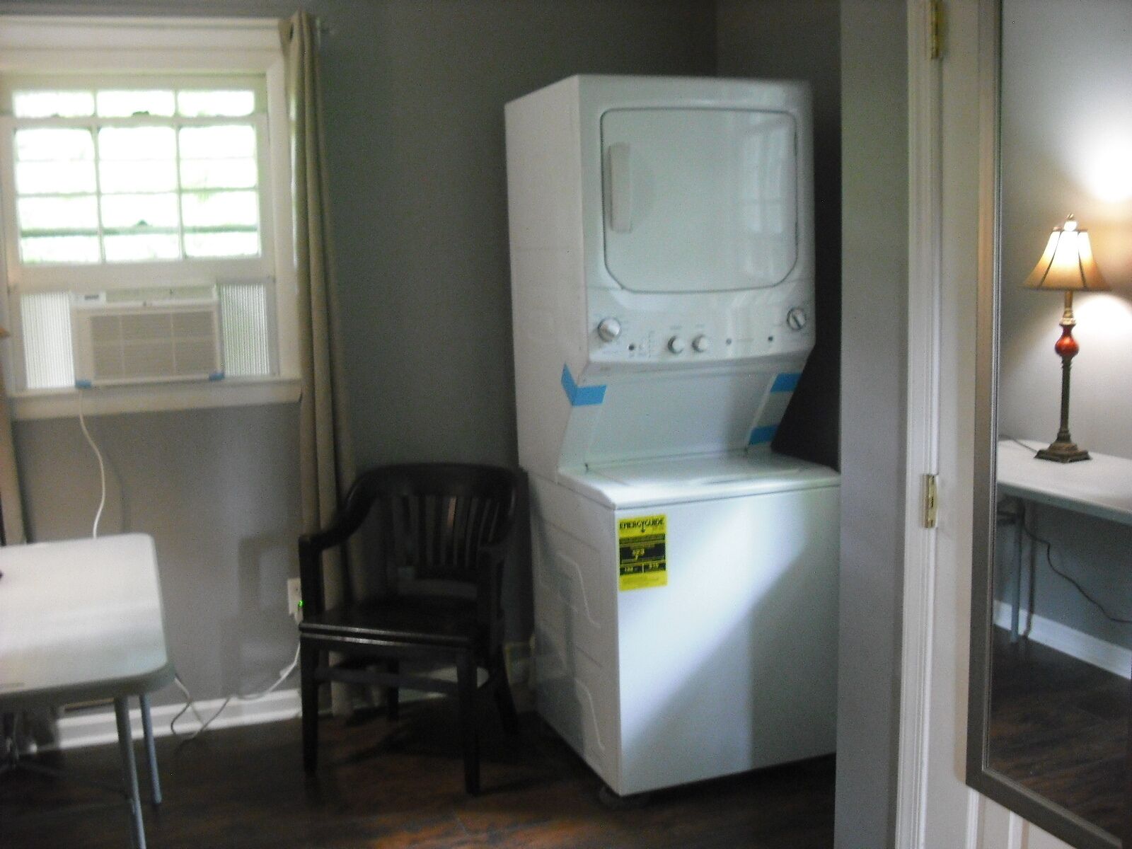 A compact laundry room with a washing machine and dryer stacked together, a chair, and a window with blinds.