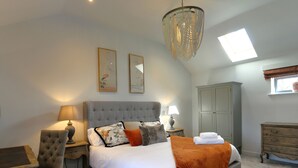 A bedroom with a tufted headboard, a large bed with white bedding, a grey upholstered chair, a wooden desk, a chandelier, and a wardrobe. - The Tyre Barn (Whitchurch)