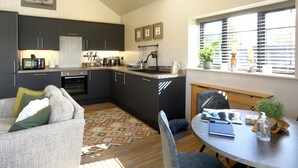 A modern kitchen with dark cabinetry, a dining area with a round table, and a large window with blinds. - The Tyre Barn (Whitchurch)