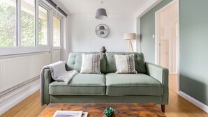 Un canapé vert agrémenté de coussins à motifs, un miroir rond, un lampadaire et une petite plante en pot, le tout disposé sur une table basse en bois, dans une pièce dotée de grandes fenêtres et d’une porte.