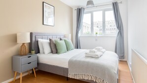 Une chambre bien rangée, avec un lit, une table de chevet, une lampe et une fenêtre avec des rideaux.