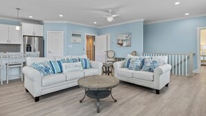Ein modernes Wohnzimmer mit zwei weißen Sofas, einem runden Couchtisch und einer Küchenzeile mit weißen Schränken und einem Edelstahlkühlschrank.