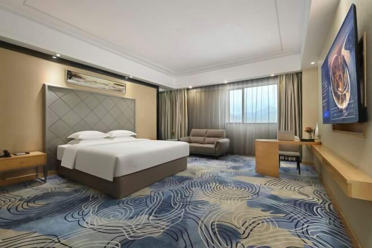 A hotel room with a bed, a desk, a chair, a sofa, and a large window with curtains.