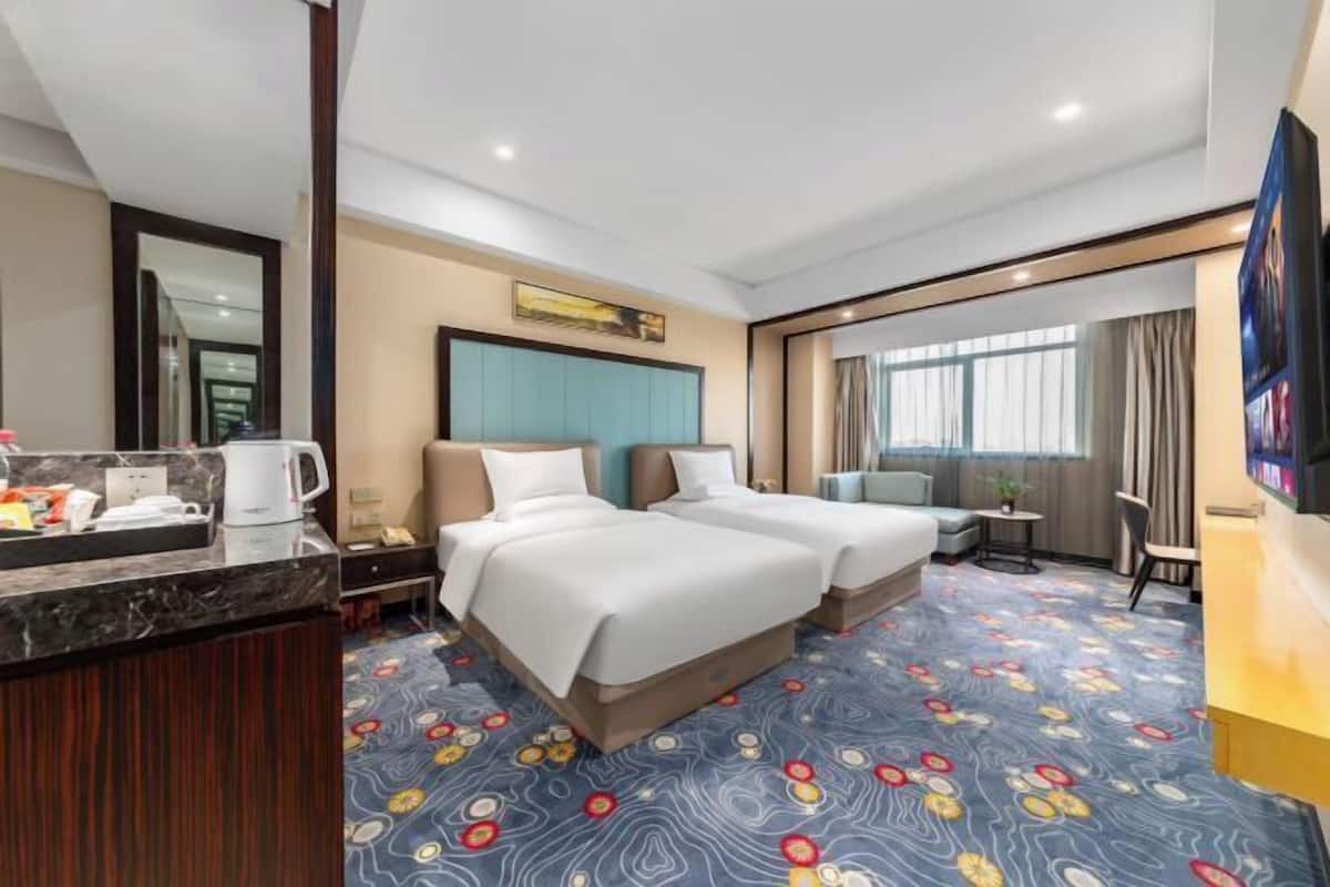 A hotel room with two beds, a desk with a coffee maker, a TV, and a large window with curtains.