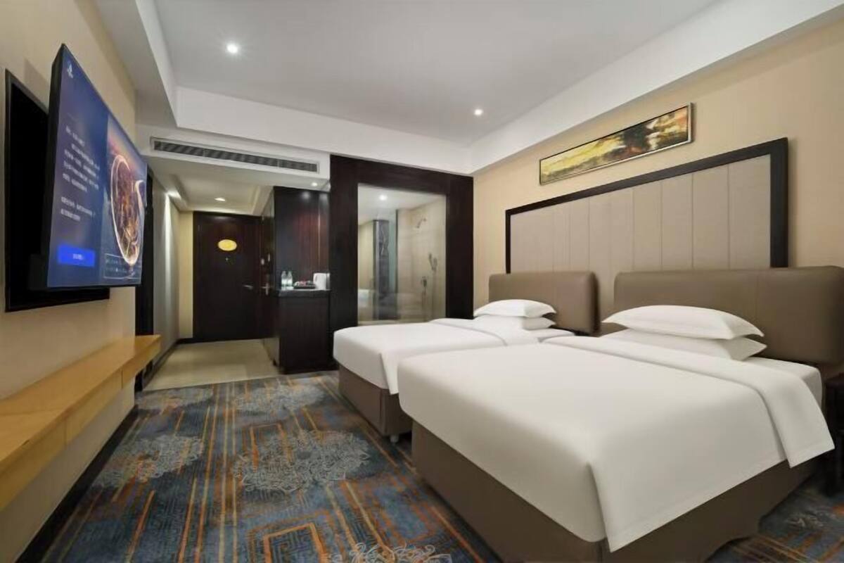 A hotel room with two beds, a TV mounted on the wall, a bathroom visible through a glass door, and a carpeted floor.