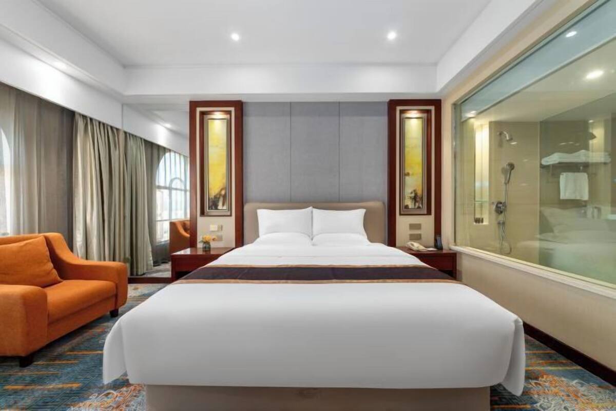 A hotel room with a large bed, a sofa, a glass shower enclosure, and a view of the city.