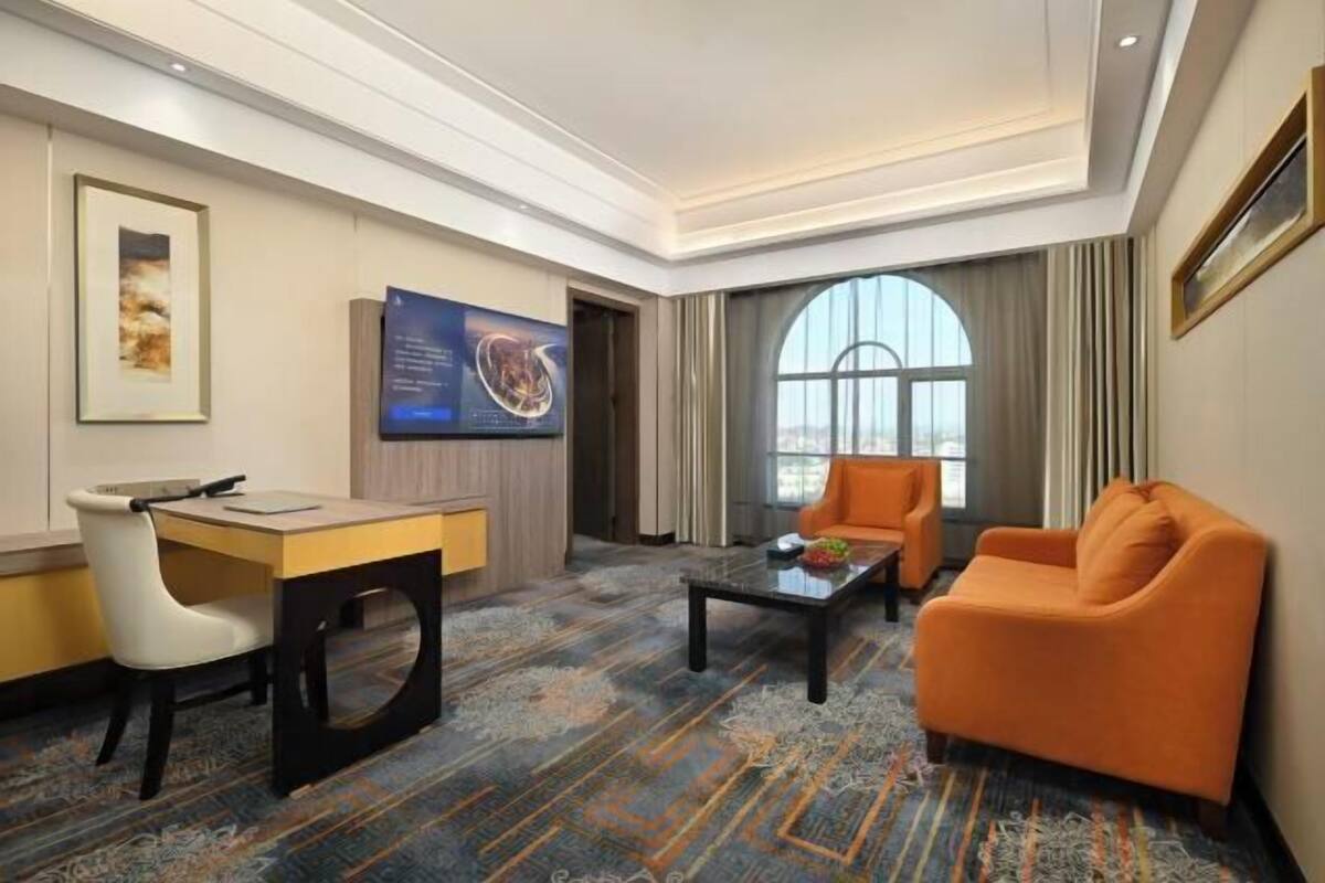 A hotel room with a desk, two chairs, a sofa, a coffee table, a large window, and artwork on the walls.