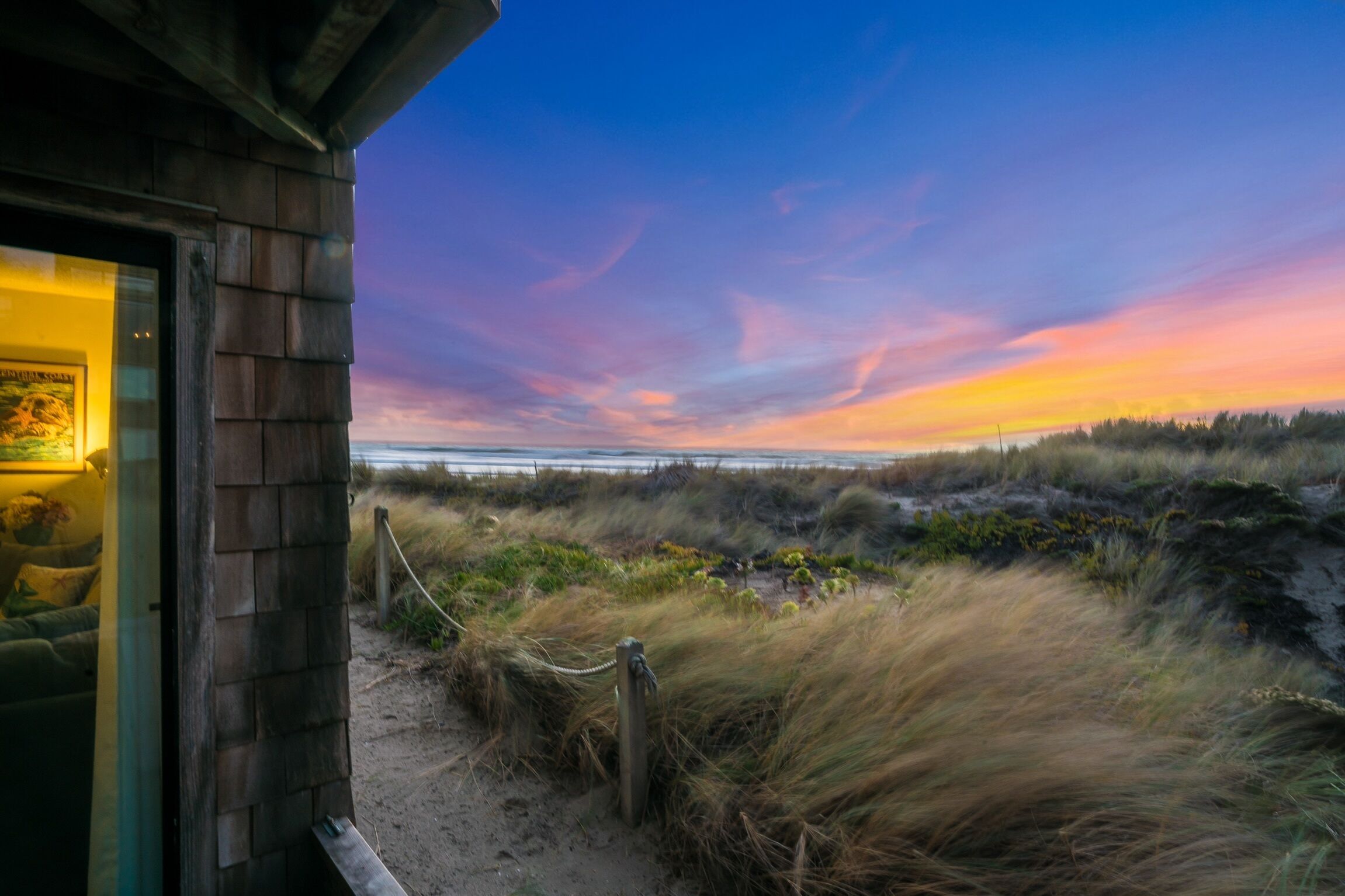 A coastal view with a wooden building, a sandy path, and a rope barrier.