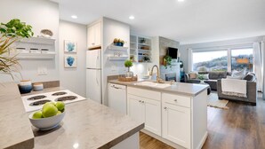 A modern kitchen with white cabinetry, a central island, and a dining area with a sofa and a TV. - Serene & Modern~ Ocean Front View~ 2 Bd 2 Bth~1st Fully Remodeled (Watsonville)