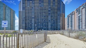 A modern high-rise building with a grid of windows and balconies, surrounded by a sandy beach and a wooden fence.