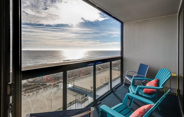 A balcony with a view of the beach and ocean. There are turquoise chairs and a small table.