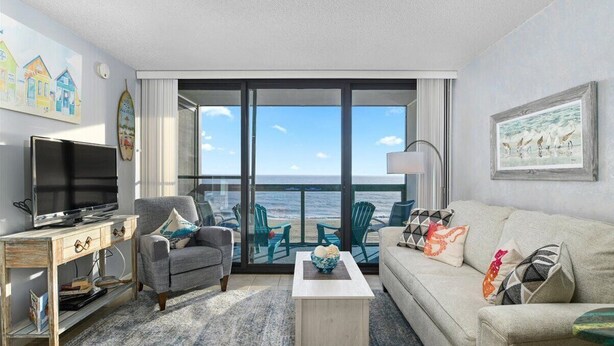 A room with a television, a sofa, a coffee table, and a view of the ocean through large windows.