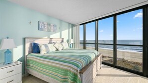 A bedroom with a large window overlooking a beach, a bed with striped bedding, and a nightstand with a lamp.