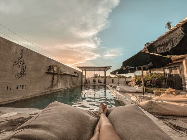 Vista de la piscina con sillas de descanso, una pérgola y una pared con el texto 'EL NIDO'.