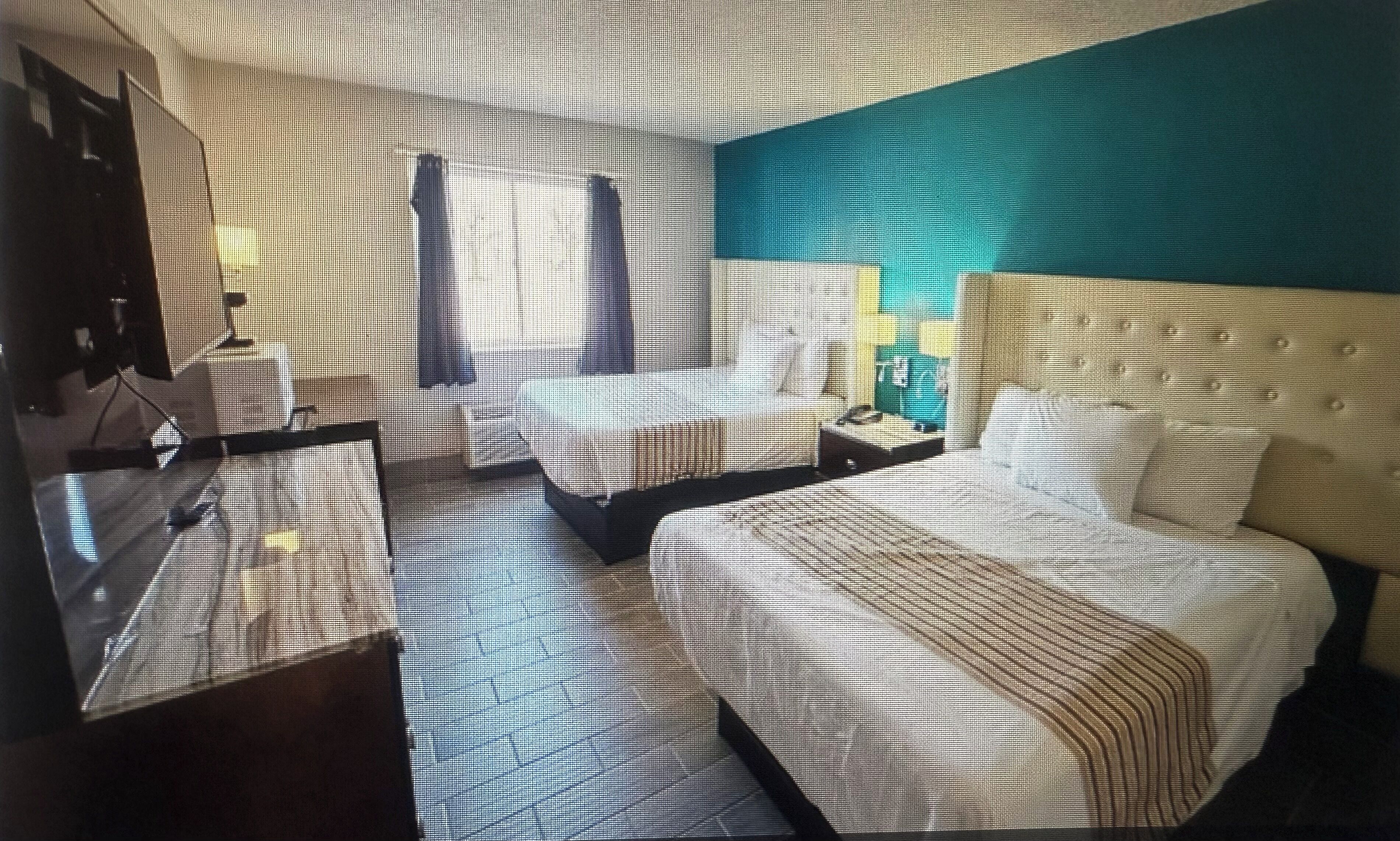 Habitación de hotel con una cama grande, un escritorio y una silla. Tiene una ventana con cortinas, un espejo y un televisor montado en la pared.