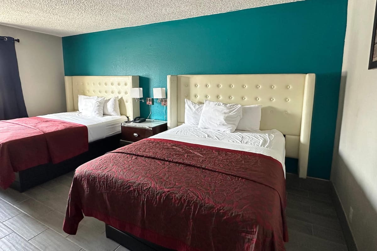 A hotel room with two beds, each with a headboard and white bedding. There are two bedside tables with lamps and a phone. The room has teal walls and a tiled floor.