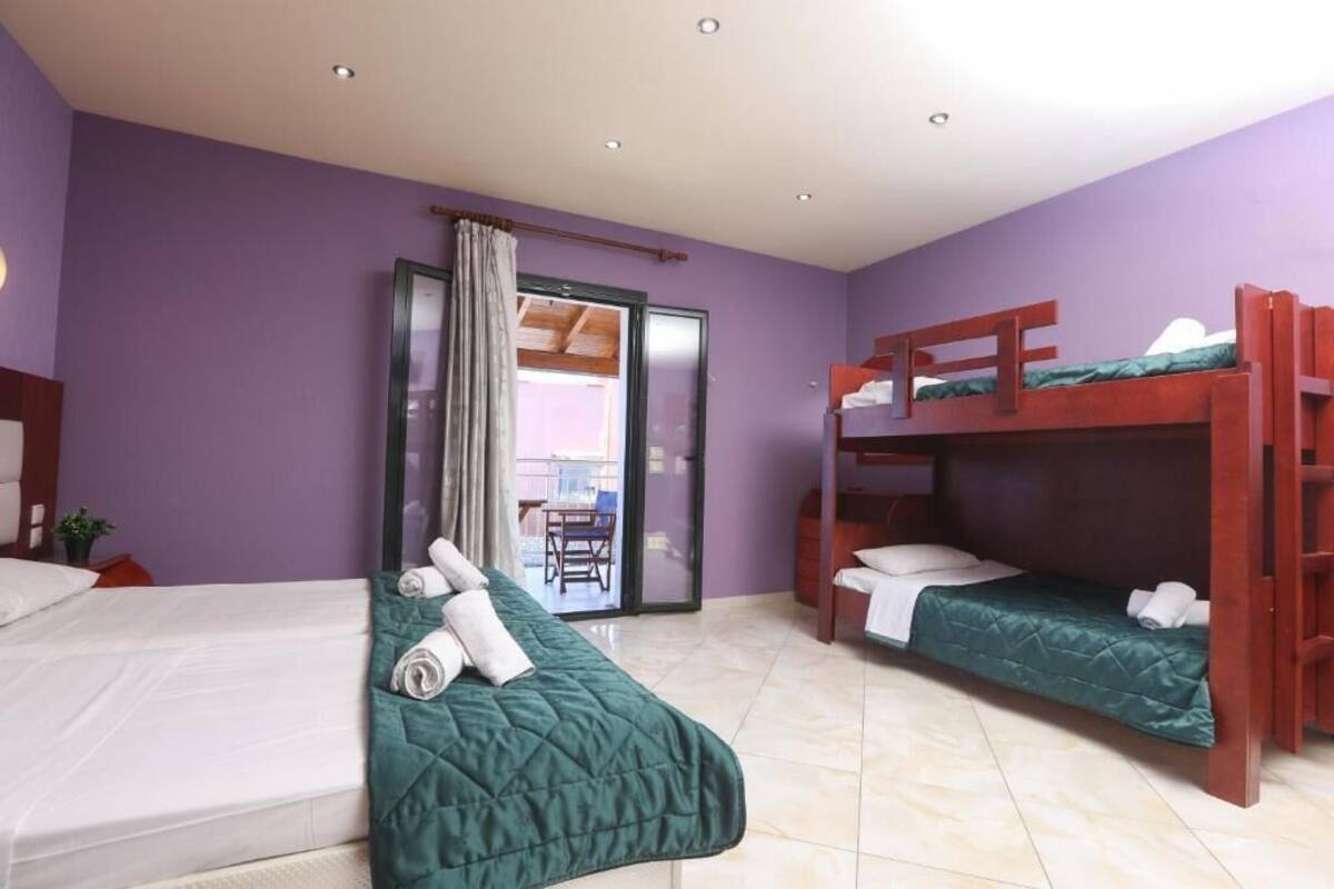 Ein Hotelzimmer mit zwei Betten, eines mit einer grünen Decke und das andere mit einem weißen Lattenrost. Rechts befindet sich ein Holzbett, und eine Glasschiebetür führt in einen Außenbereich.