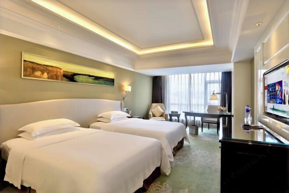 A hotel room with two beds, a desk, a chair, a TV, and a painting on the wall.