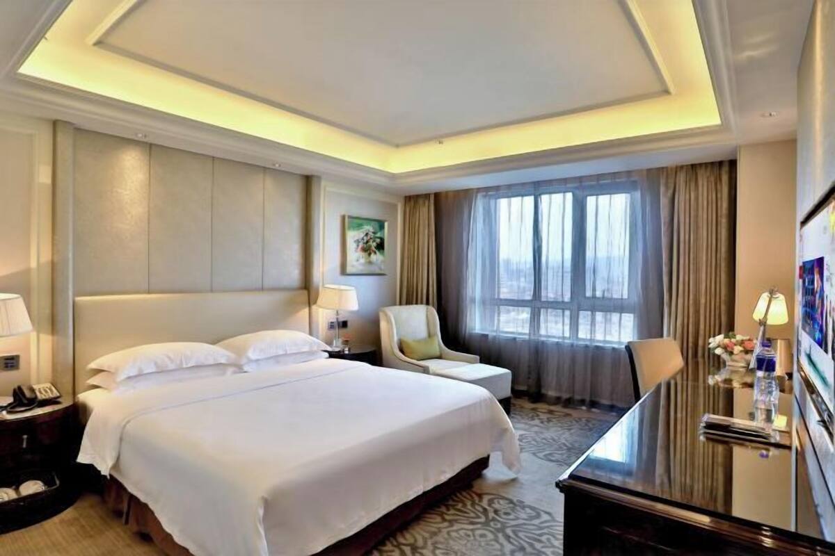 A hotel room with a large bed, a desk, a chair, a lamp, and a painting on the wall.