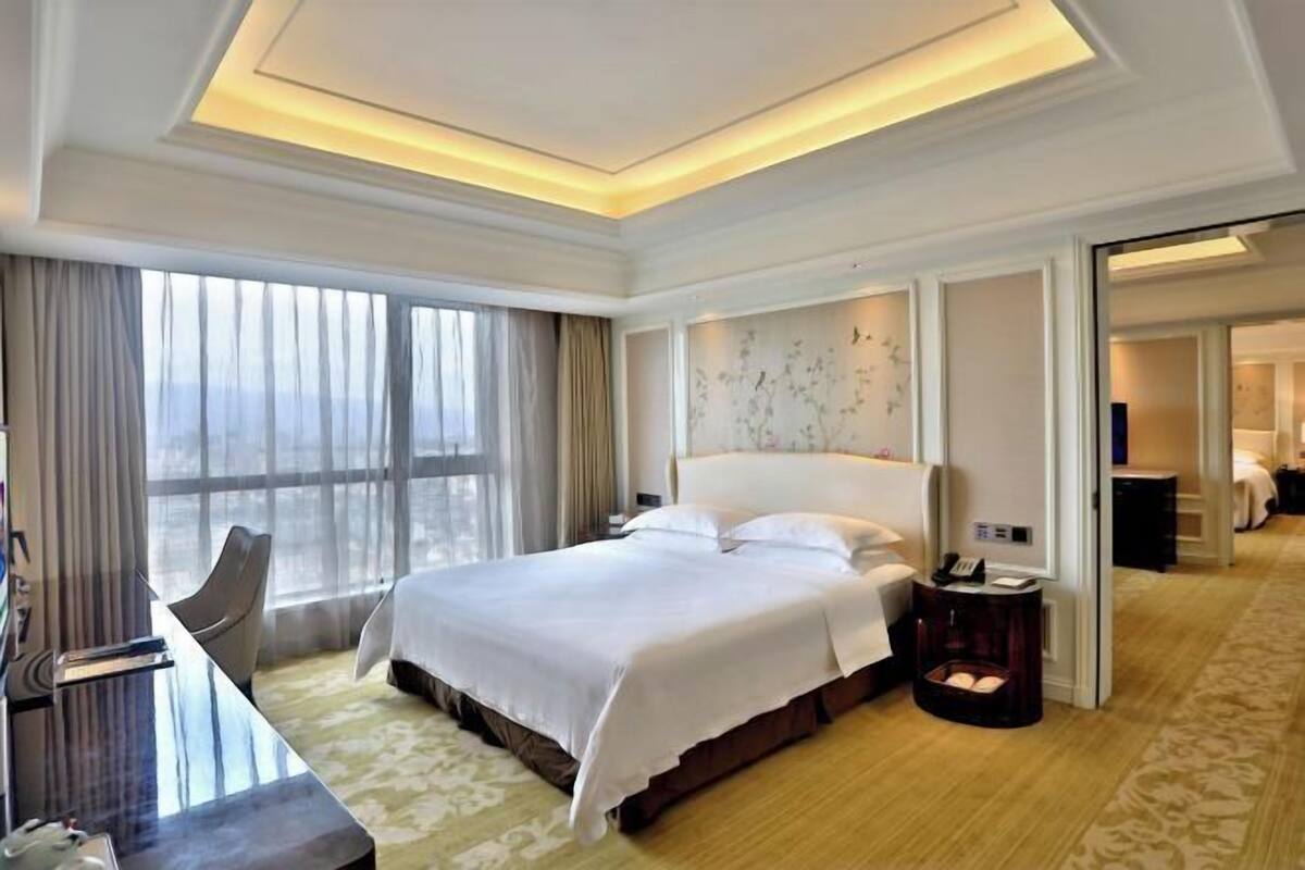 A hotel room with a large bed, a desk, and a chair. There is a window with curtains, a wall-mounted phone, and a small table with a lamp.