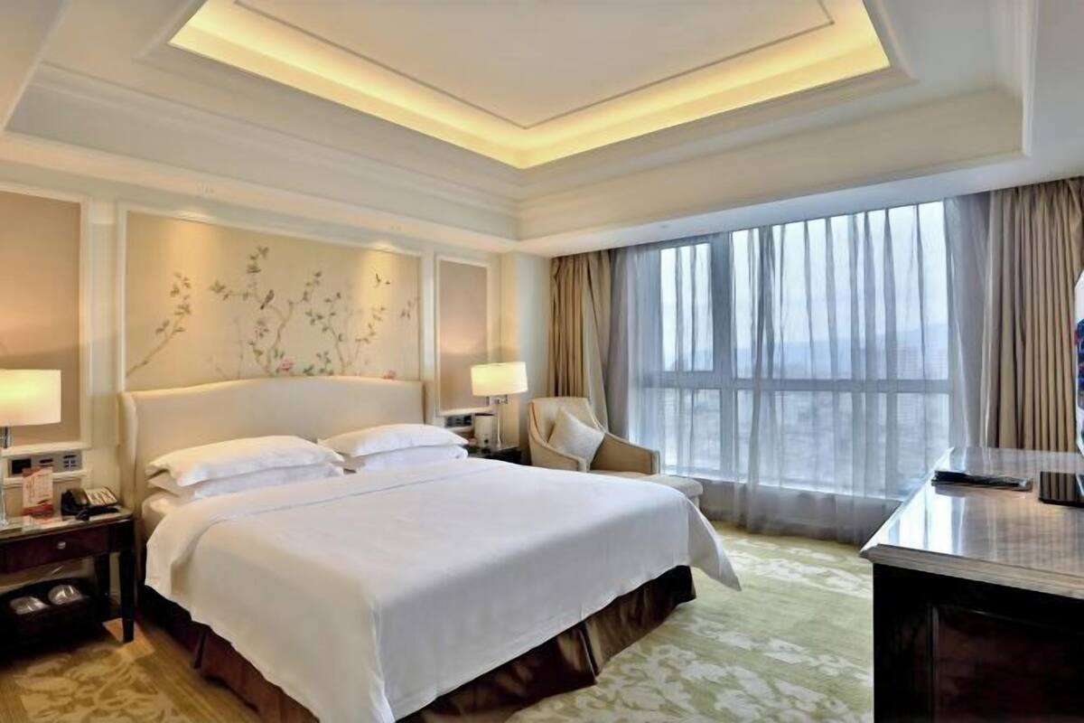 A hotel room with a large bed, a desk, a chair, and a window with curtains.