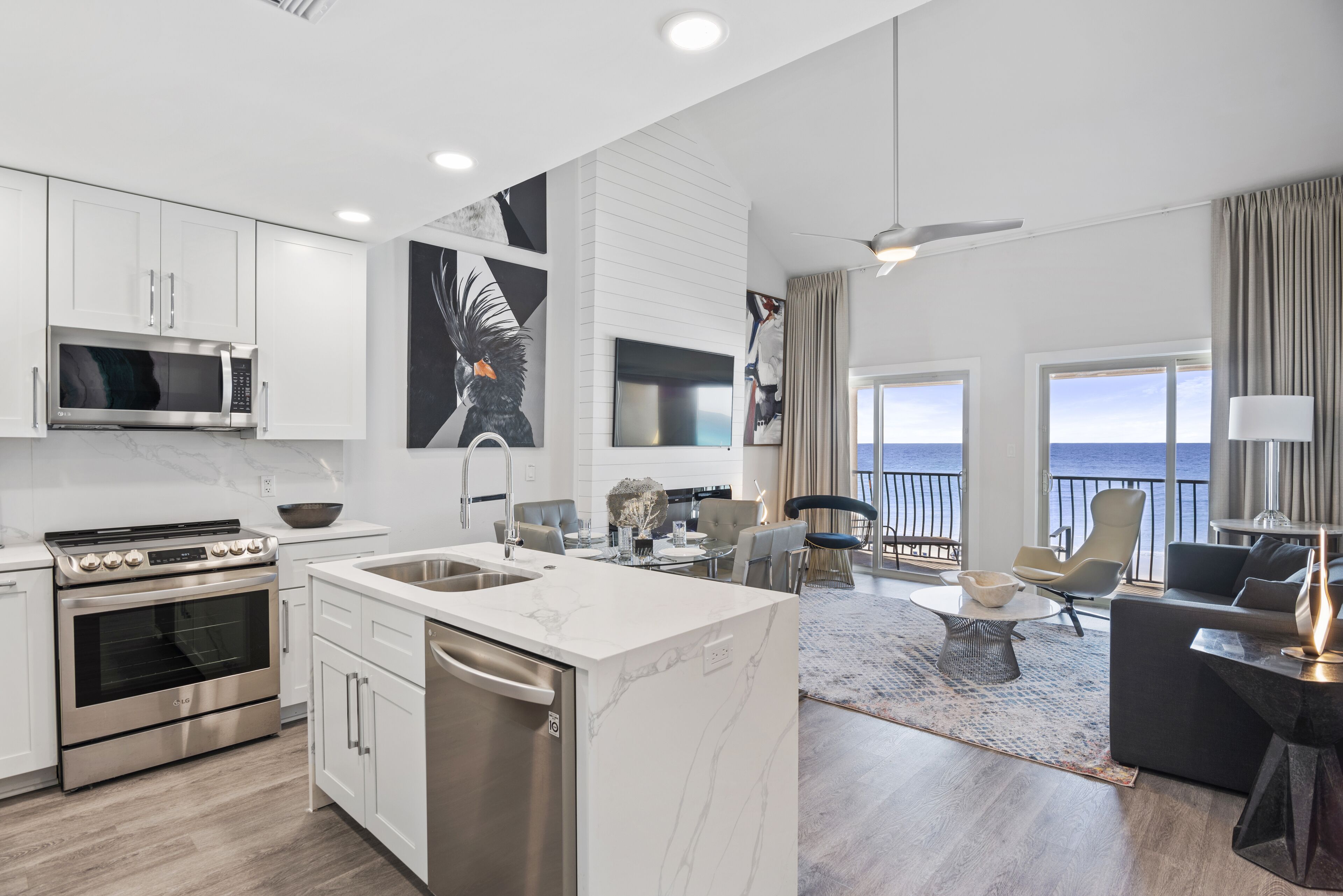 A modern kitchen with a built-in oven and microwave, a dining area with a view of the sea, and a living room with a large window and a ceiling fan.