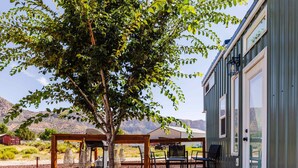 A wooden deck with outdoor furniture, a tree, and a building with large windows. - Zion Tiny Home • Pet Friendly • Stargazing Deck (Apple Valley)
