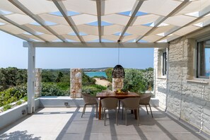 A covered outdoor dining area with a table and chairs, overlooking a scenic landscape with a view of the sea and hills.