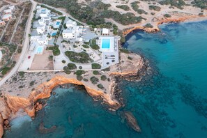 An aerial view of a coastal resort with multiple buildings, a swimming pool, and rocky cliffs.