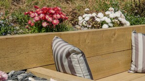 A wooden bench with striped cushions, surrounded by a garden with red and white flowers. - 14 Parc Delfryn (Brynteg)