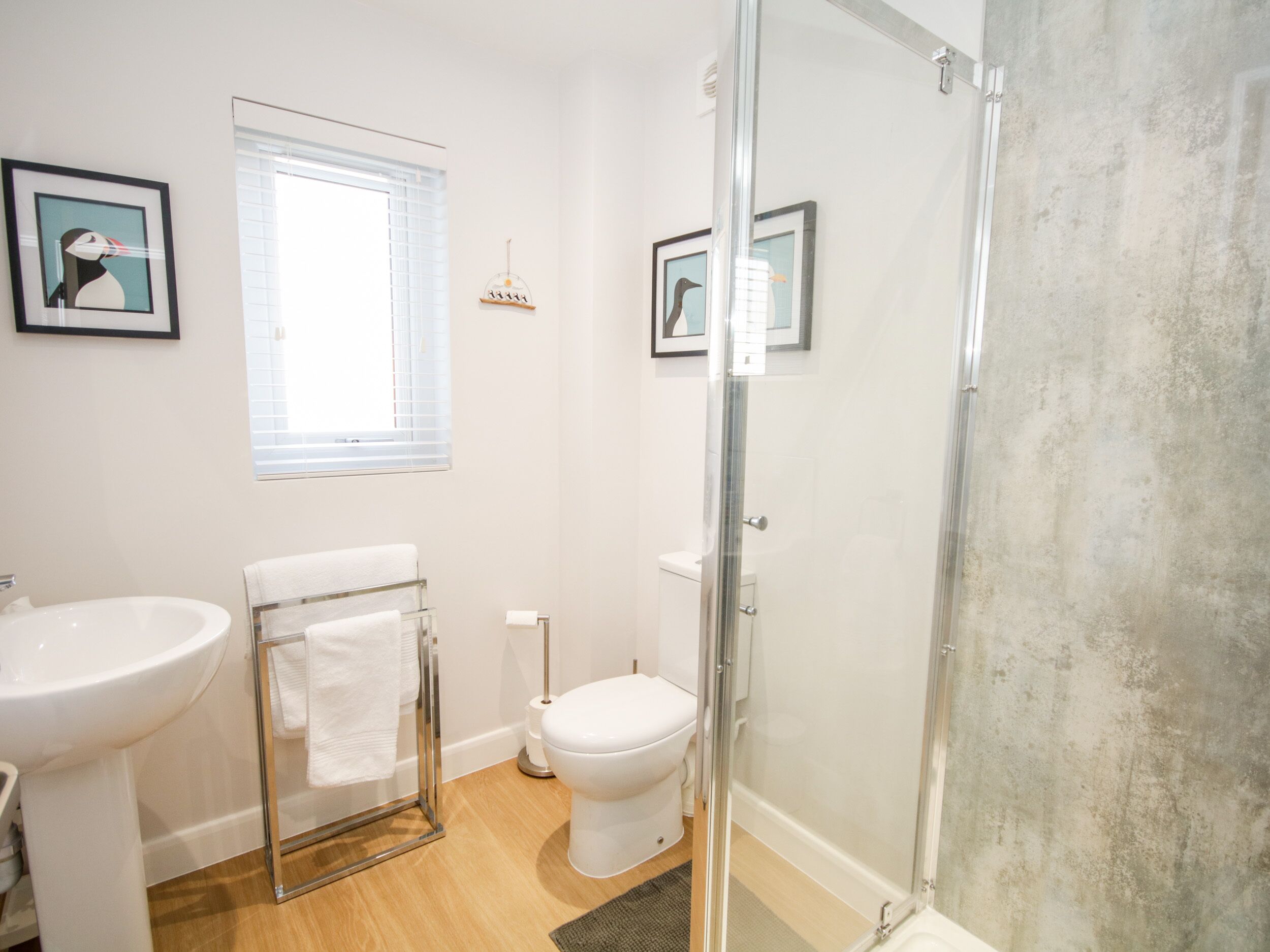 A modern bathroom with a glass-enclosed shower, a white sink, and a toilet. There are framed pictures on the walls and a window with blinds.