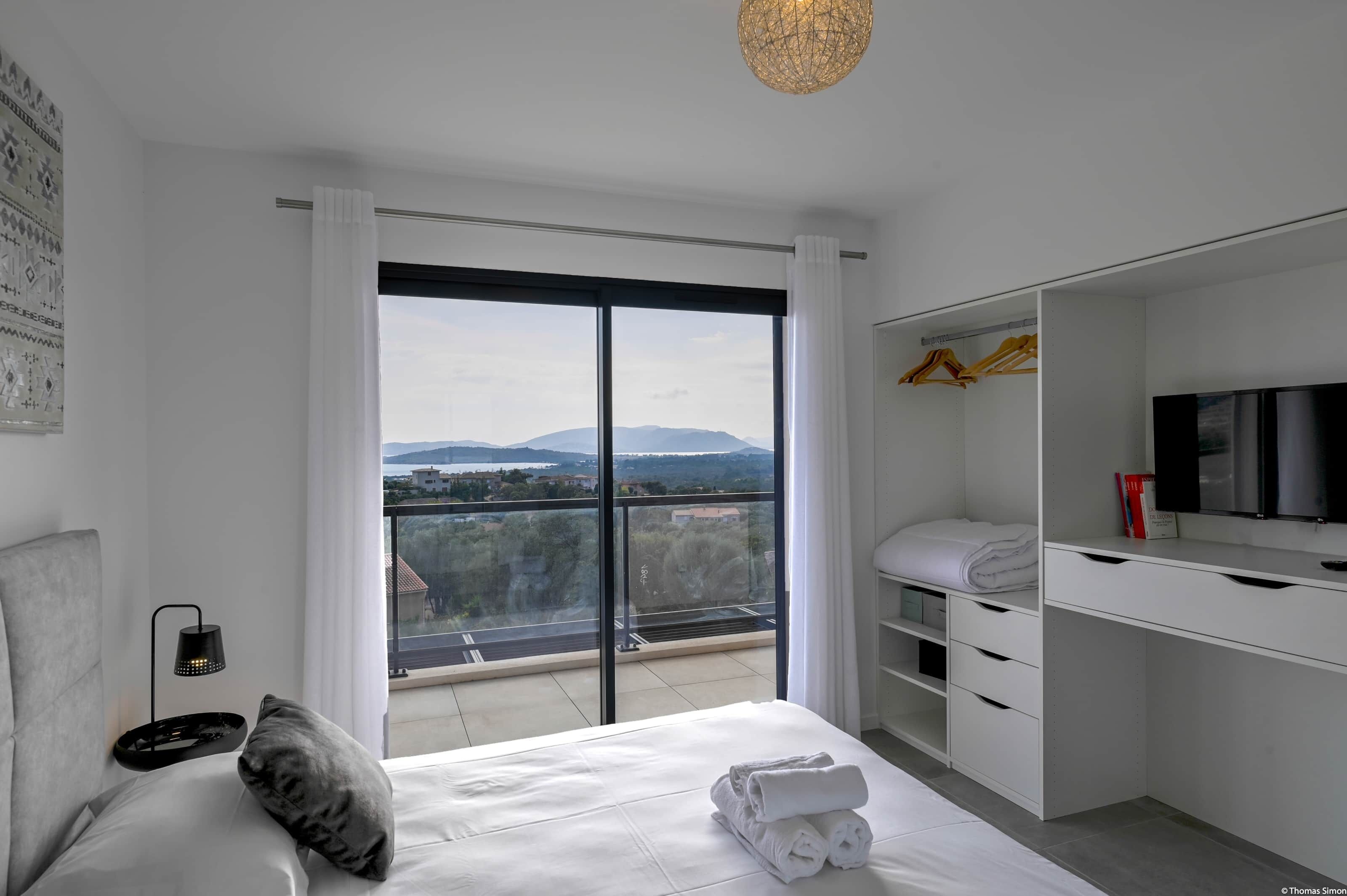 A modern hotel room with a large bed, a desk, a TV, and a view of the city and mountains.