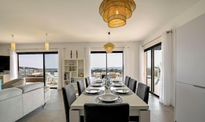 A modern dining area with a large table set for six, a sofa, and a view of the city through large windows.