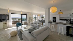Ein modernes Wohnzimmer mit Sofa, Hocker und Essbereich. Es gibt eine Küche mit Bar und einem großen Fenster mit Blick auf die Stadt.