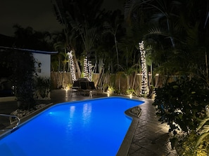 A swimming pool surrounded by palm trees and a wooden fence, with string lights along the fence.