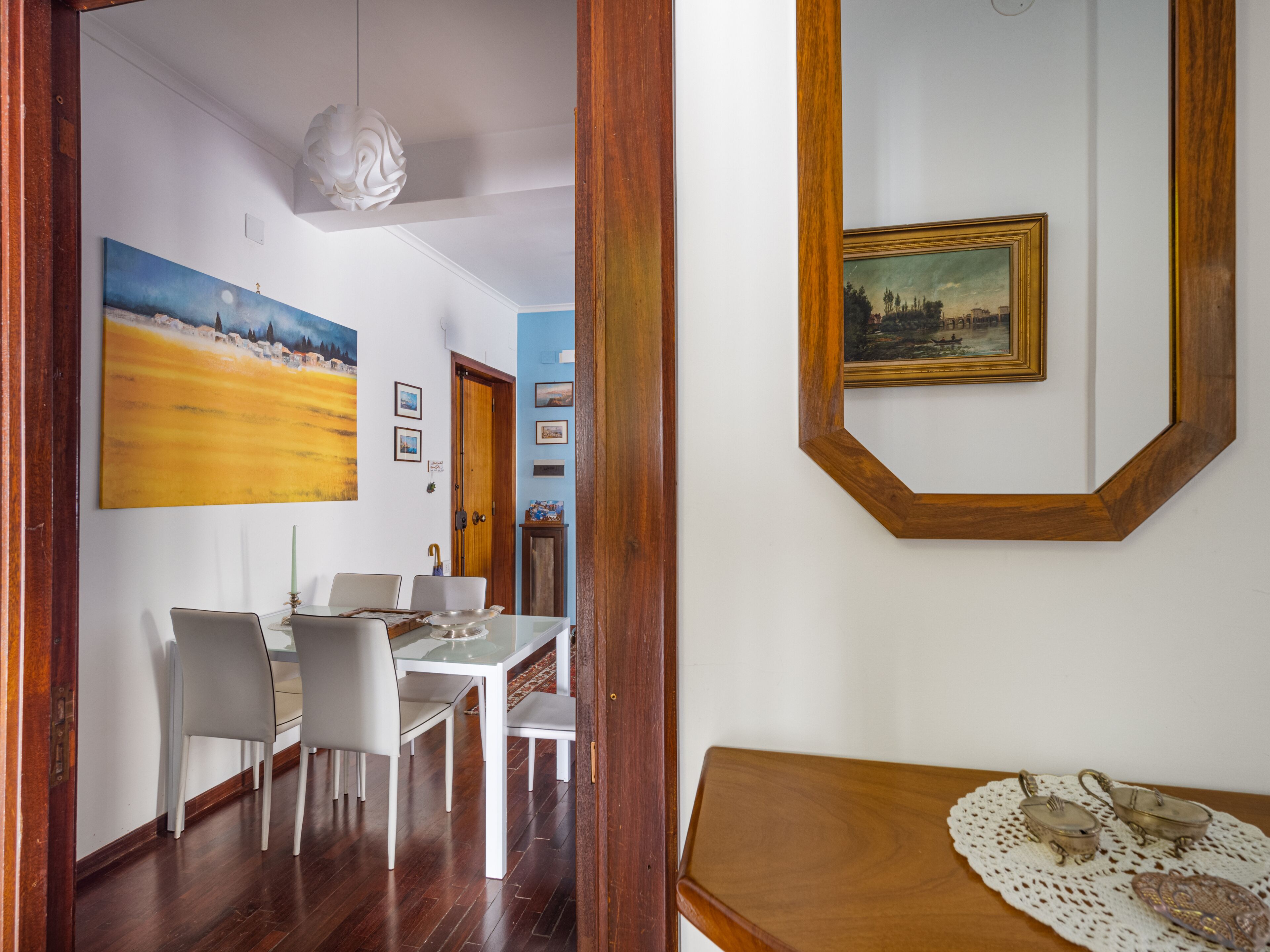 Un comedor con una mesa y sillas blancas, un cuadro en la pared y un aparador de madera con objetos decorativos.
