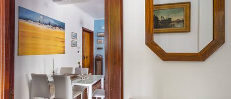 Uma área de refeições com uma mesa e cadeiras brancas, um quadro na parede e um aparador de madeira com objetos decorativos.