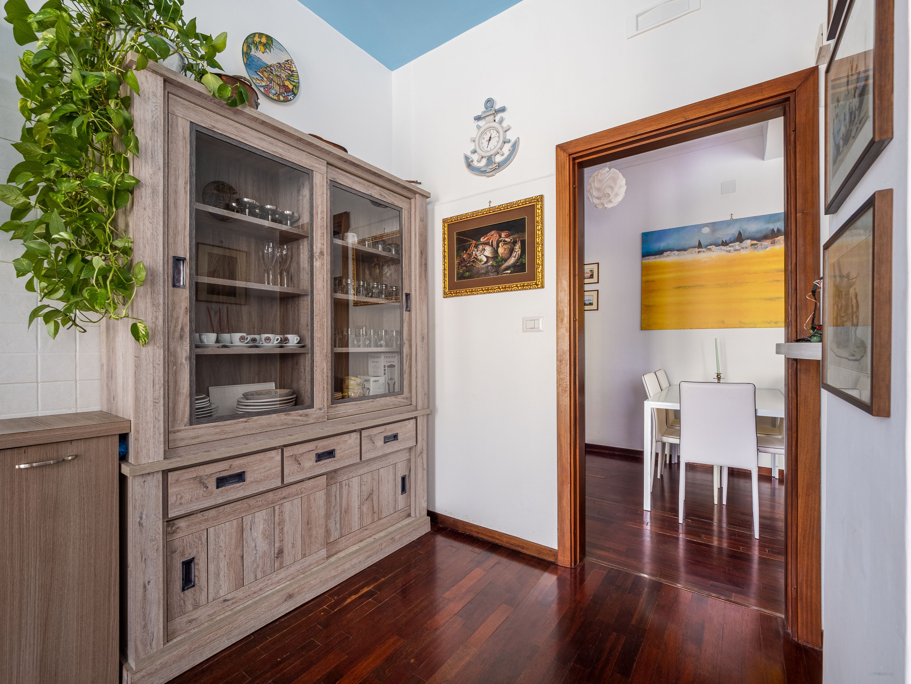 Una stanza con un armadietto in legno contenente bicchieri e stoviglie, un tavolo da pranzo con sedie e una porta che dà su un'altra stanza.