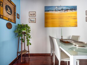 Uma sala de jantar com mesa de vidro, cadeiras brancas, planta em vaso e um quadro de paisagem.
