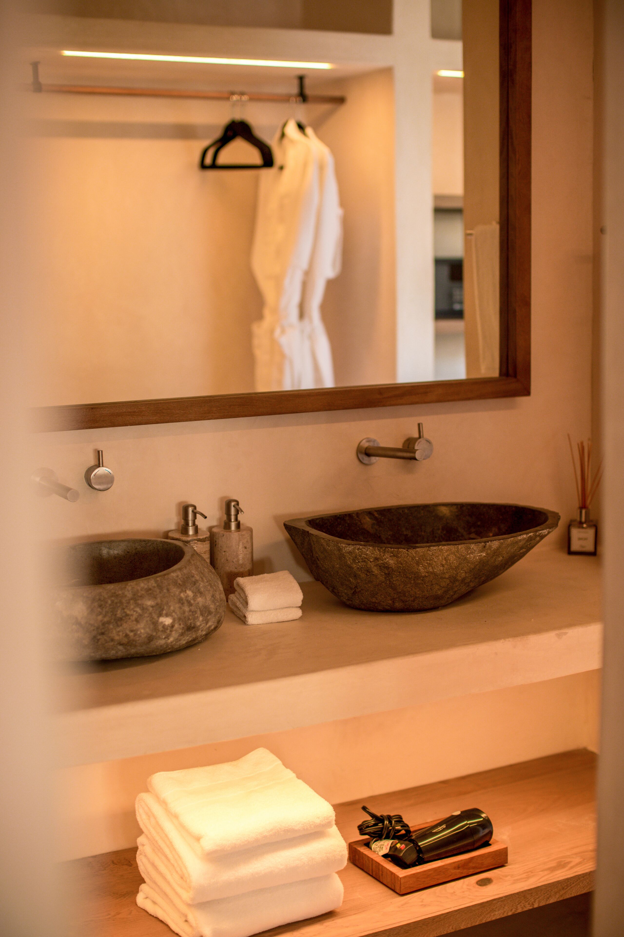 A bathroom with a large mirror, two sinks, a towel rack, and a wooden shelf with towels and a bottle.