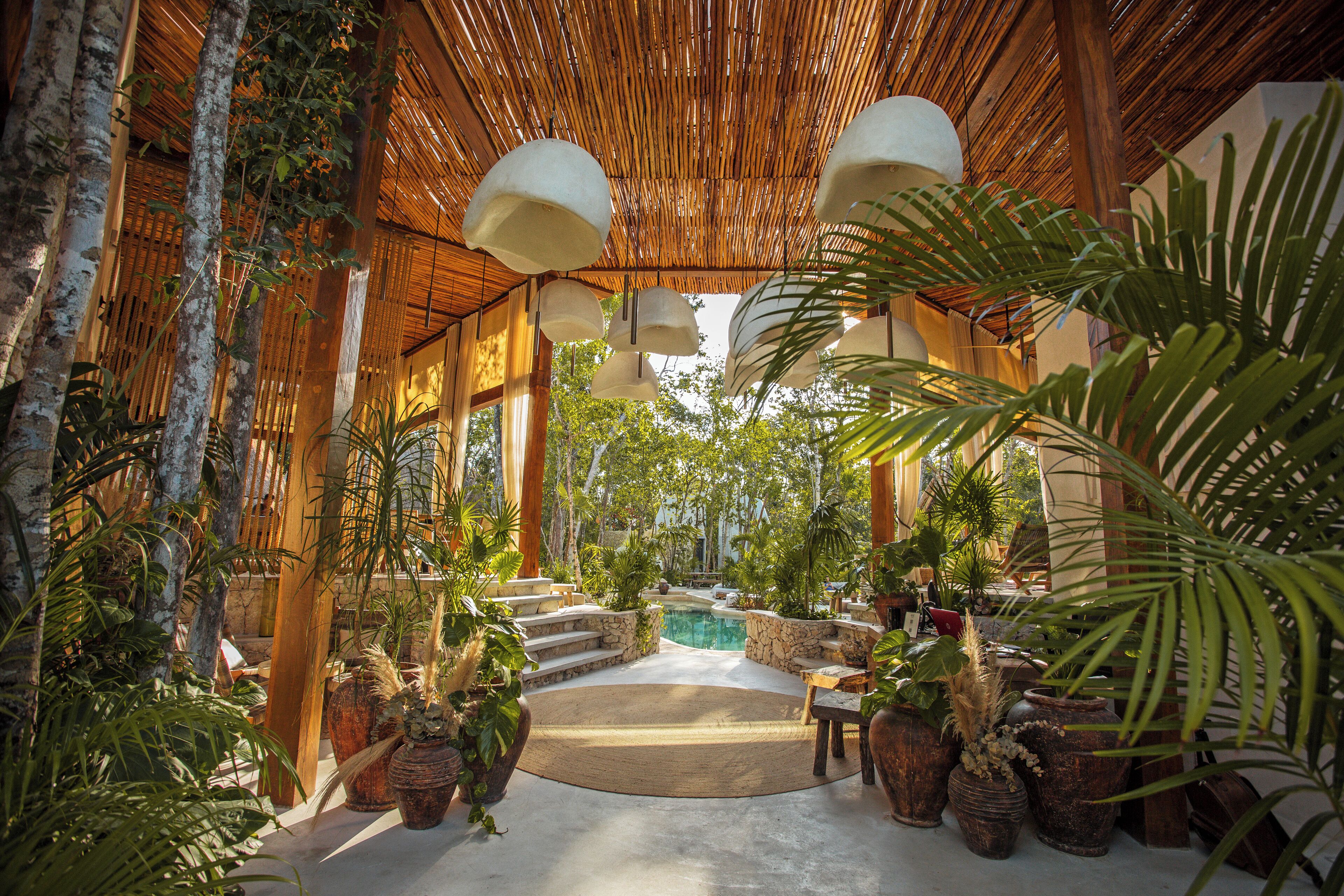 A covered outdoor area with a pool, surrounded by potted plants and wooden furniture.