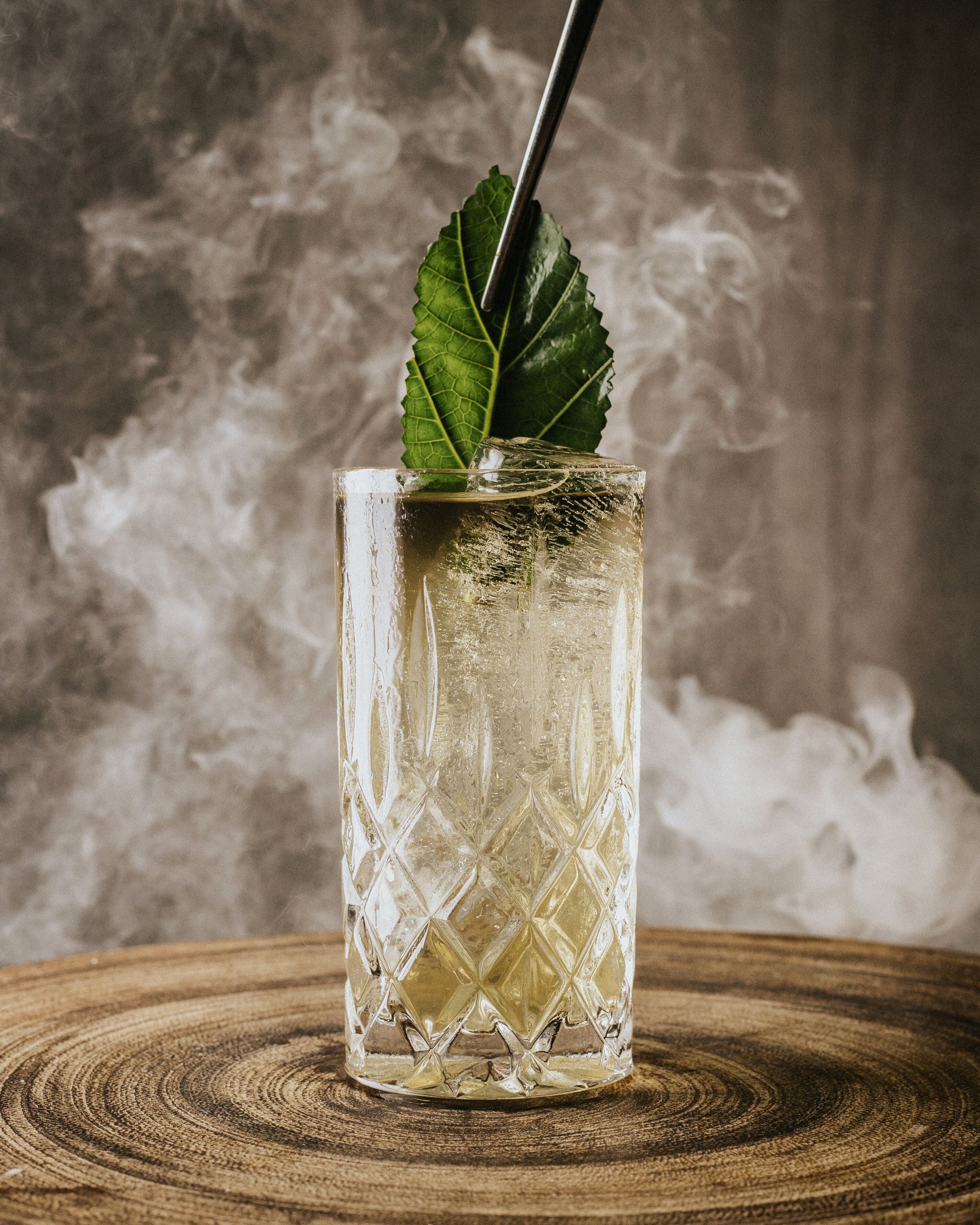 A clear glass with a diamond pattern, a green leaf garnish, and a straw, placed on a wooden surface with a textured background.