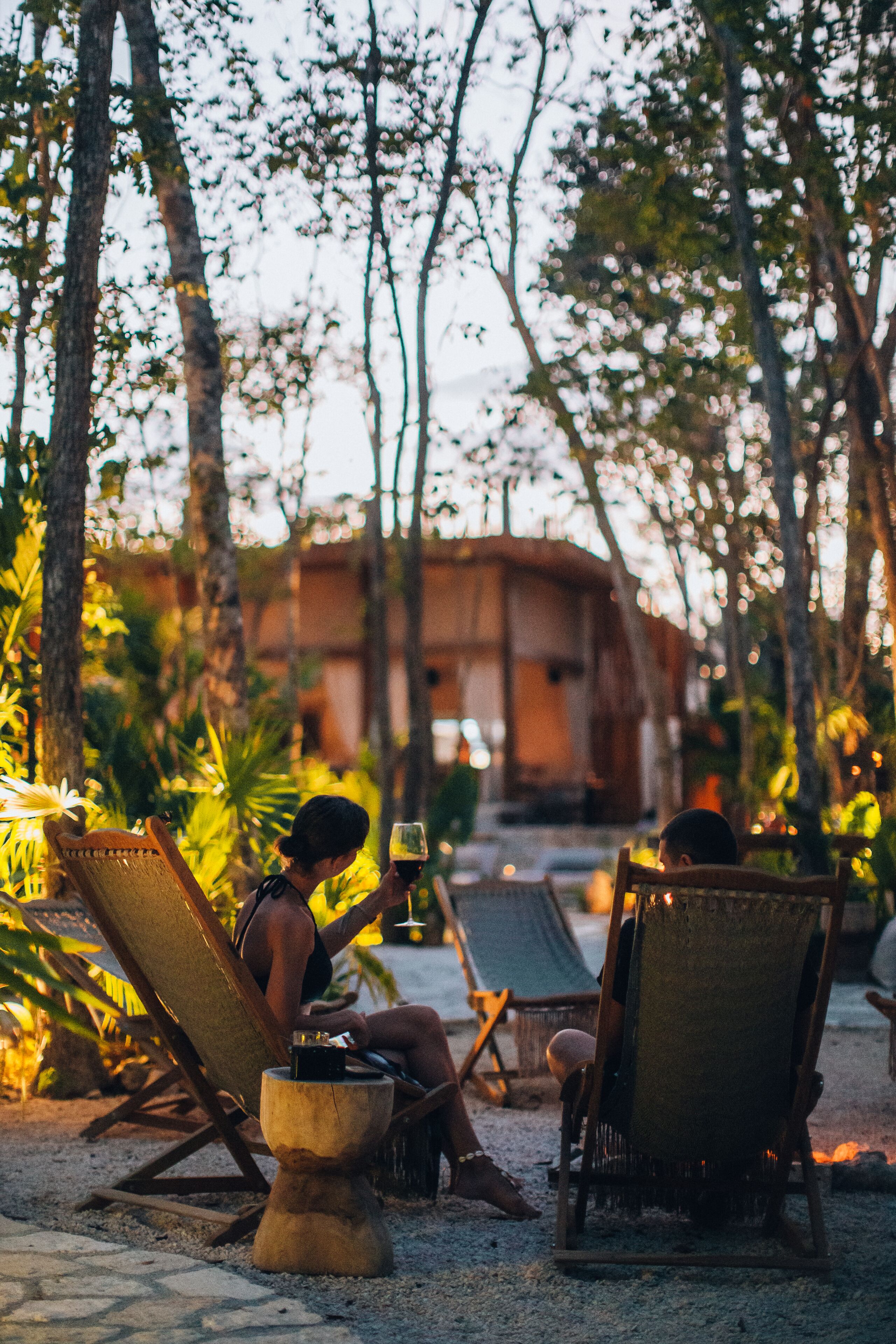 Two lounge chairs with a small table between them, surrounded by trees and a building in the background.