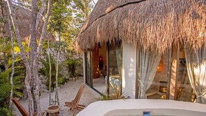 Una estructura con techo de paja, puerta de vidrio, jacuzzi y muebles de madera en un jardín.
