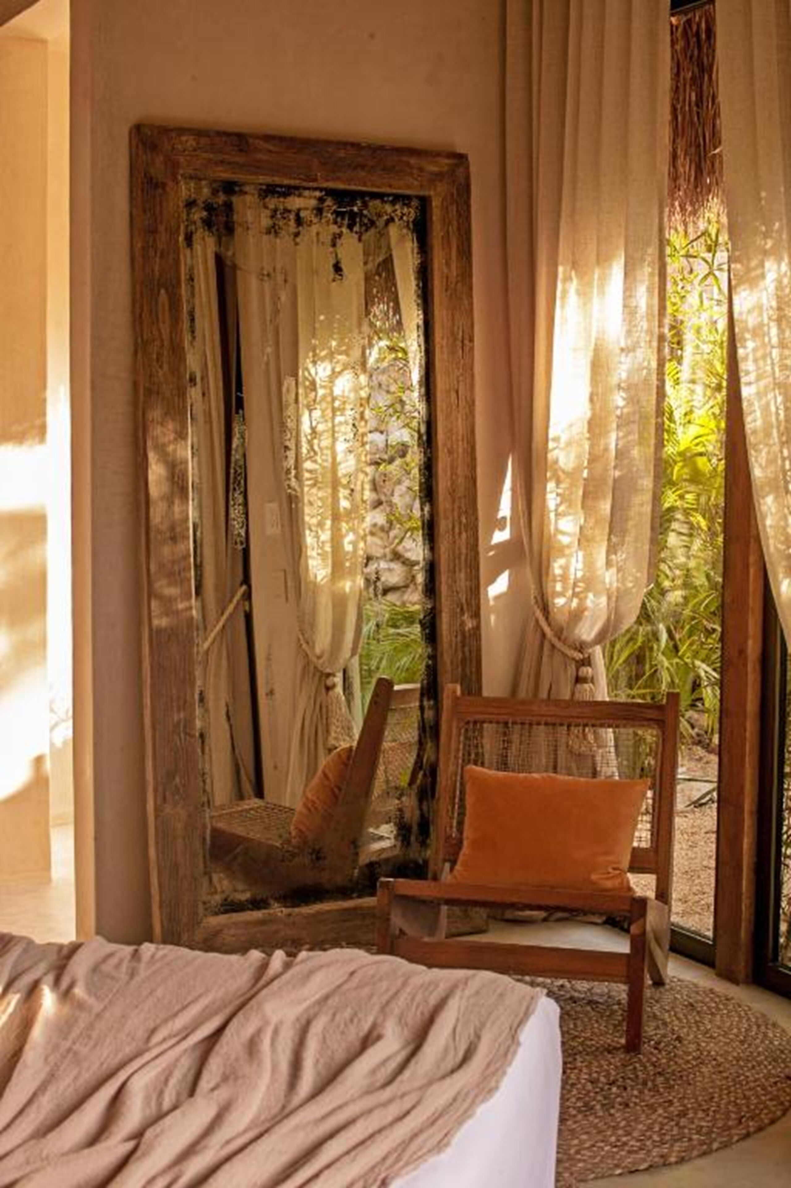 Un dormitorio con cama, una silla de madera con cojín naranja y un ventanal con cortinas traslúcidas.