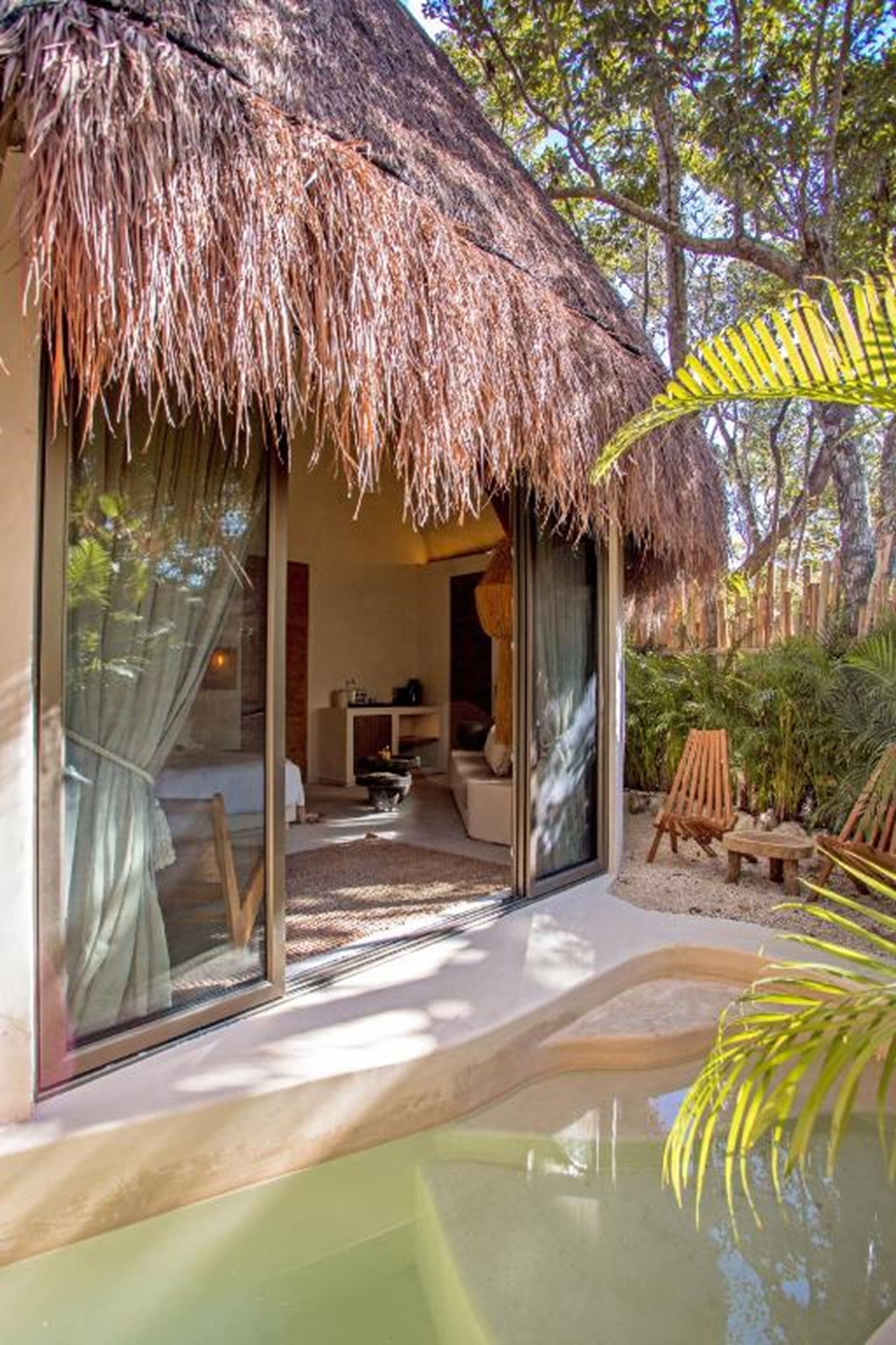 Una estructura con techo de paja y una puerta de cristal que da a una habitación con una cama, una mesita y una silla.