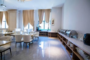 Un comedor moderno con mobiliario blanco, una estación de café y ventanas amplias con cortinas.